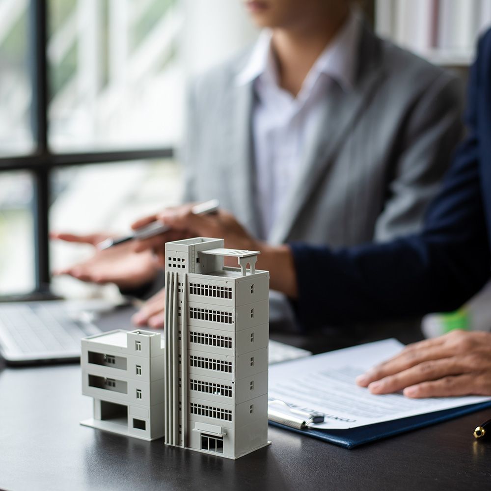 Modell eines mehrstöckigen Gebäudes auf einem Tisch vor zwei Personen in Businesskleidung, die Dokumente und Laptop nutzen