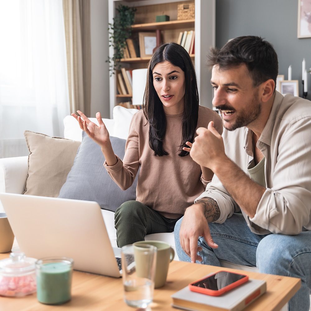 Zwei Personen sitzen auf einem Sofa vor einem Laptop auf einem Tisch mit Tassen und einem Smartphone