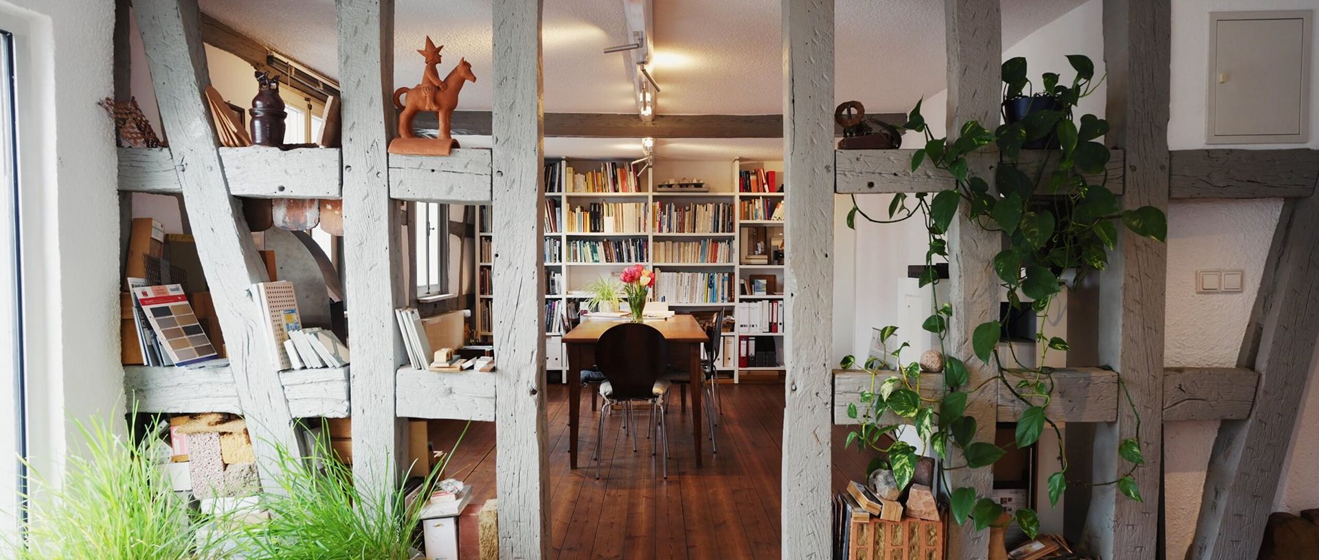 Wohnzimmer mit Holzbalken, Bücherregalen, Esstisch mit Stühlen und Pflanzen